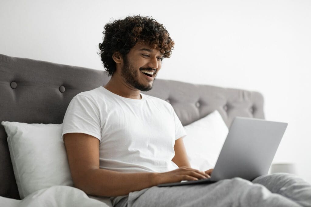 Young indian man typing on laptop, working in bed